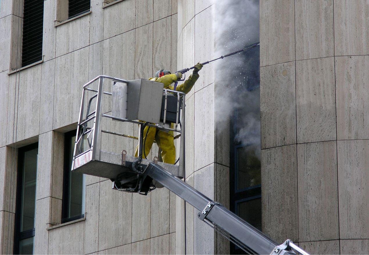 Arbeiter in gelbem Schutzanzug bei der Fassadenreinigung mit Hochdruckreiniger auf einer Arbeitsbühne – Arbeitsbühne mieten für sichere Reinigung in großer Höhe